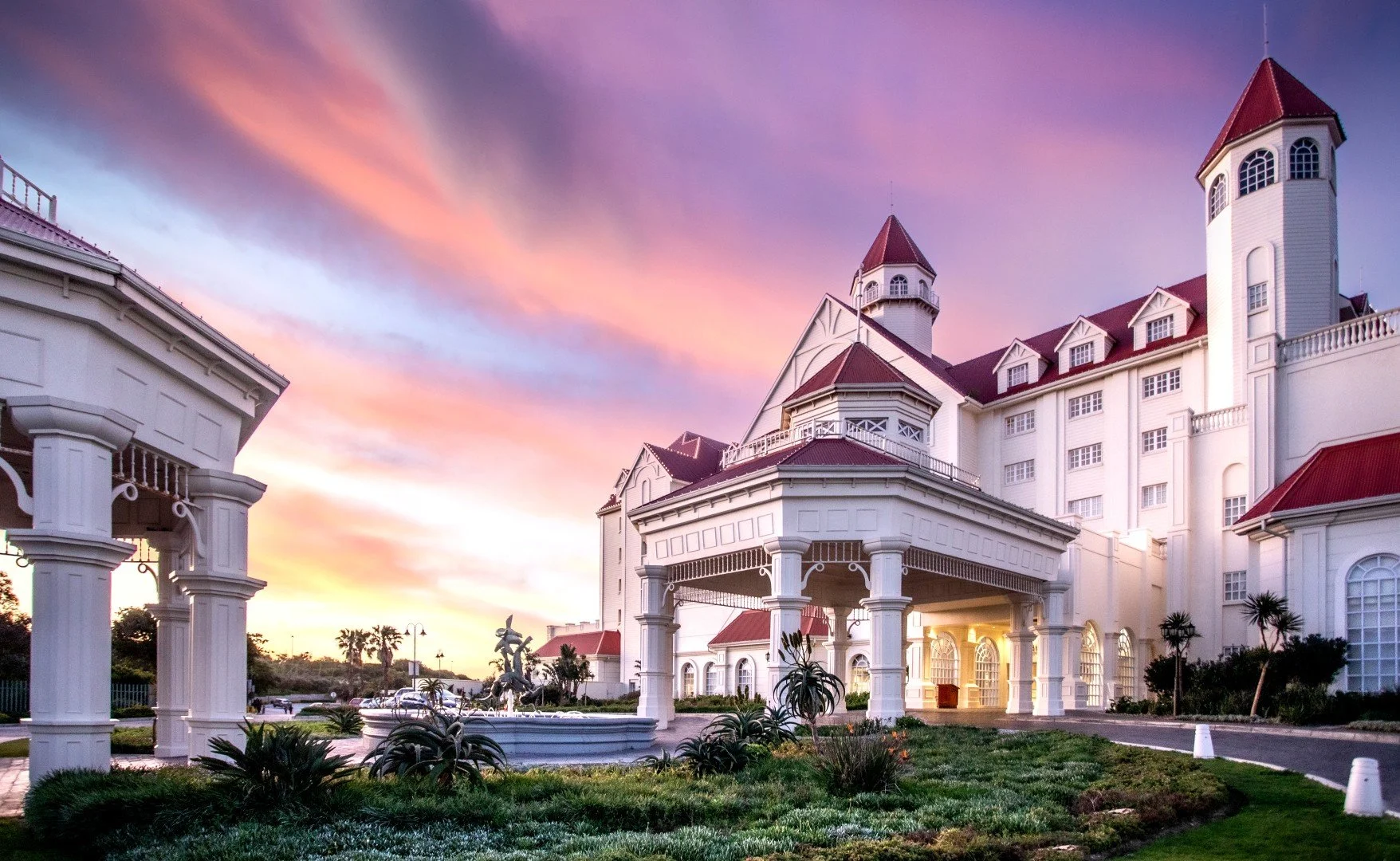 A grand, white hotel with red roofs and multiple turrets at sunset. The foreground features a fountain and manicured gardens, with a colorful sky in the background.