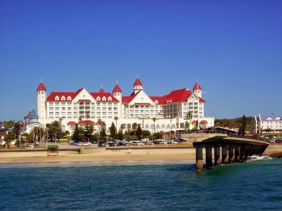 A large white hotel with red roofs and turrets, situated along a coastline with a beach and a pier extending into the water.