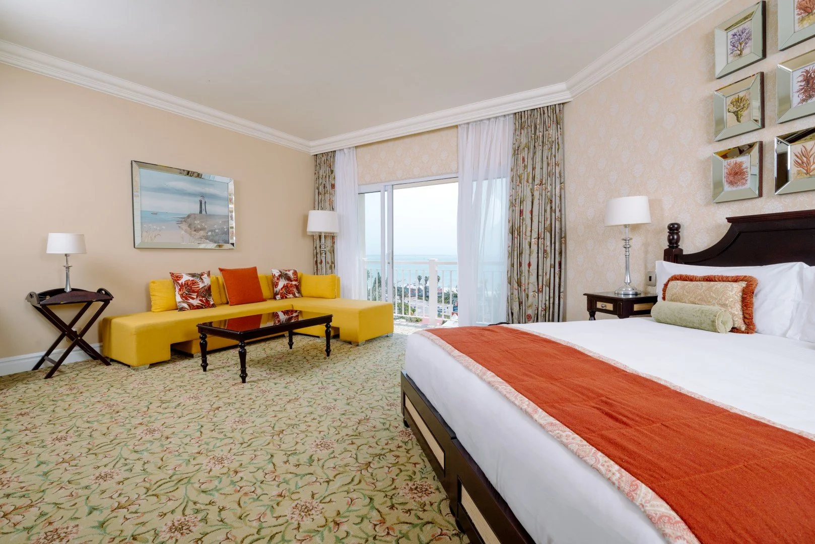 Hotel room with a large bed, colorful pillows, a yellow sofa, and a balcony view of the ocean.