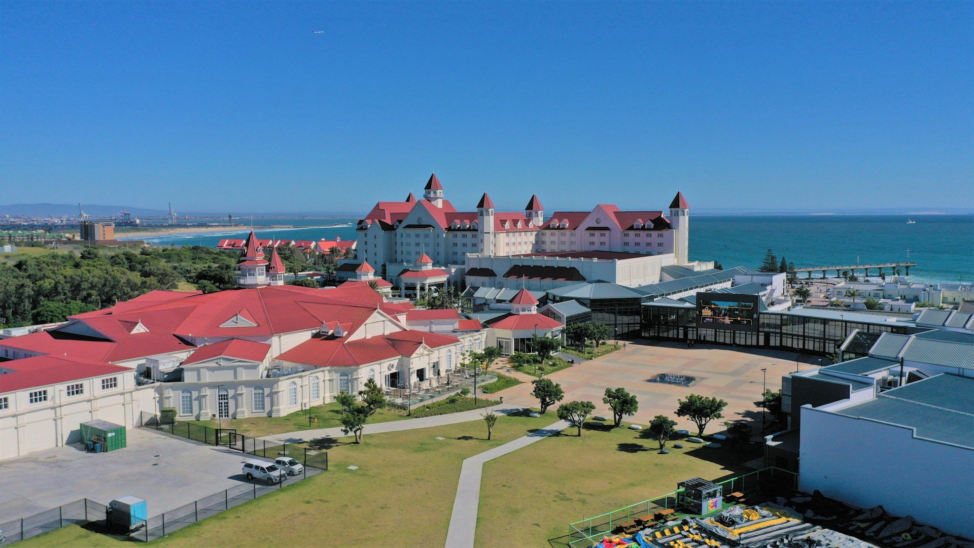 A large coastal hotel with white walls and red roofs, overlooking the ocean with a pier in the background. There are green trees and open spaces in the foreground, and a clear blue sky above.
