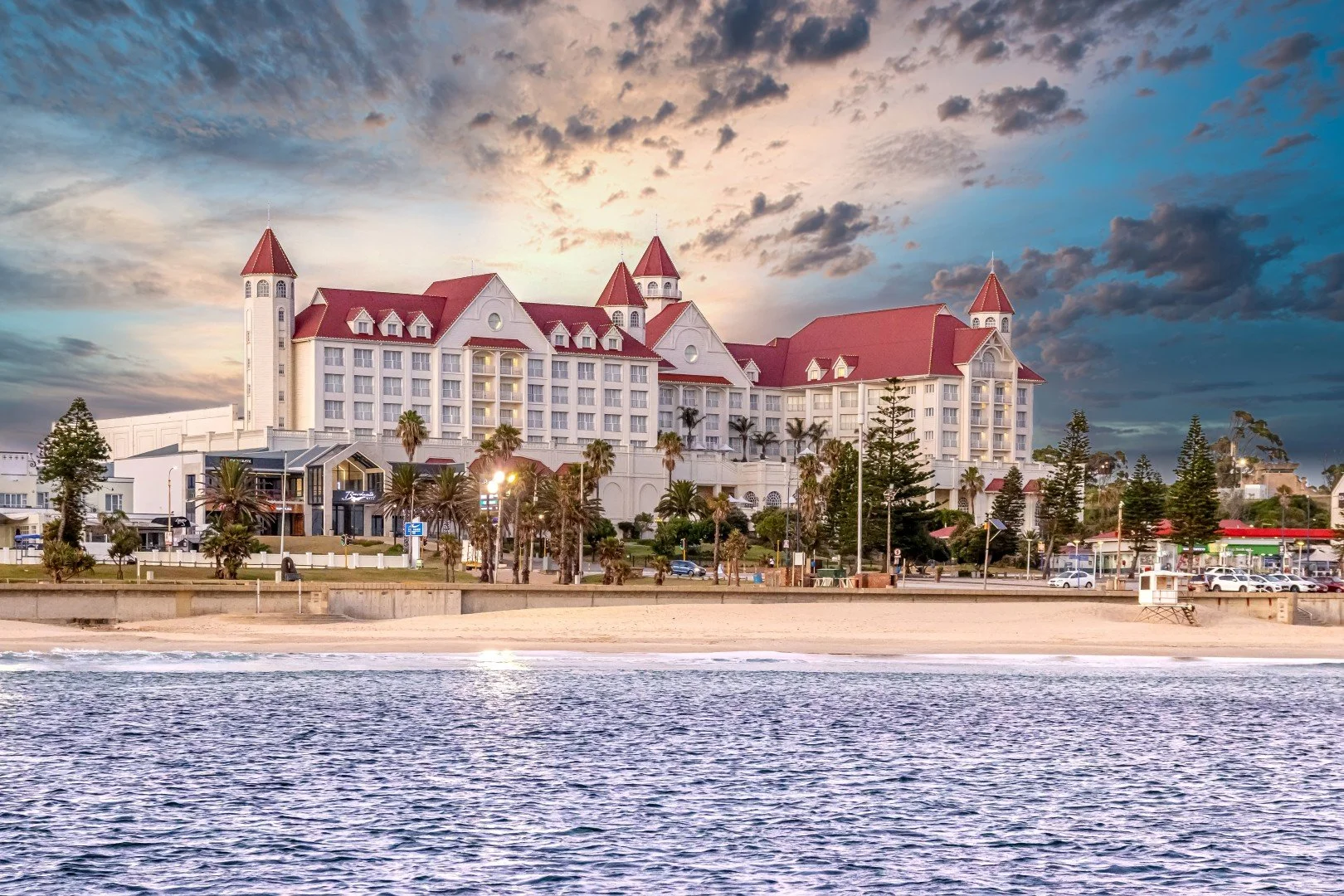 A large white hotel with red rooftops by the beach during sunset, with palm trees and a parking lot in front.