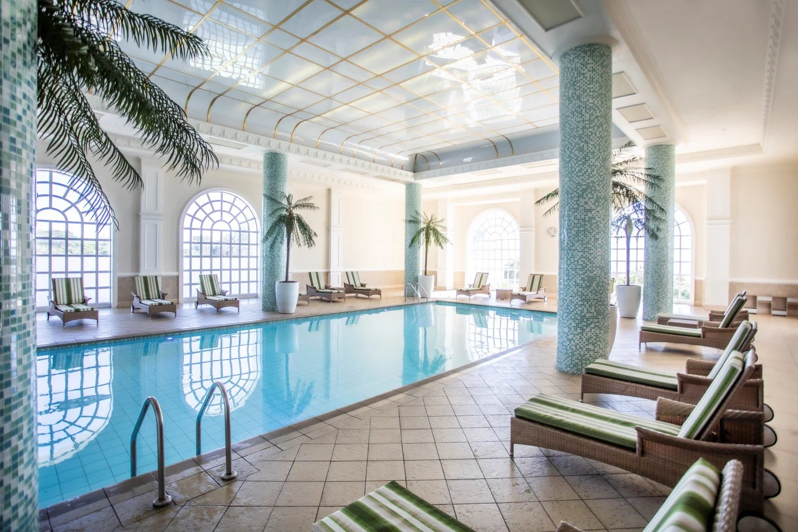 Indoor swimming pool with lounge chairs, palm trees in white pots, and large arched windows allowing natural light.