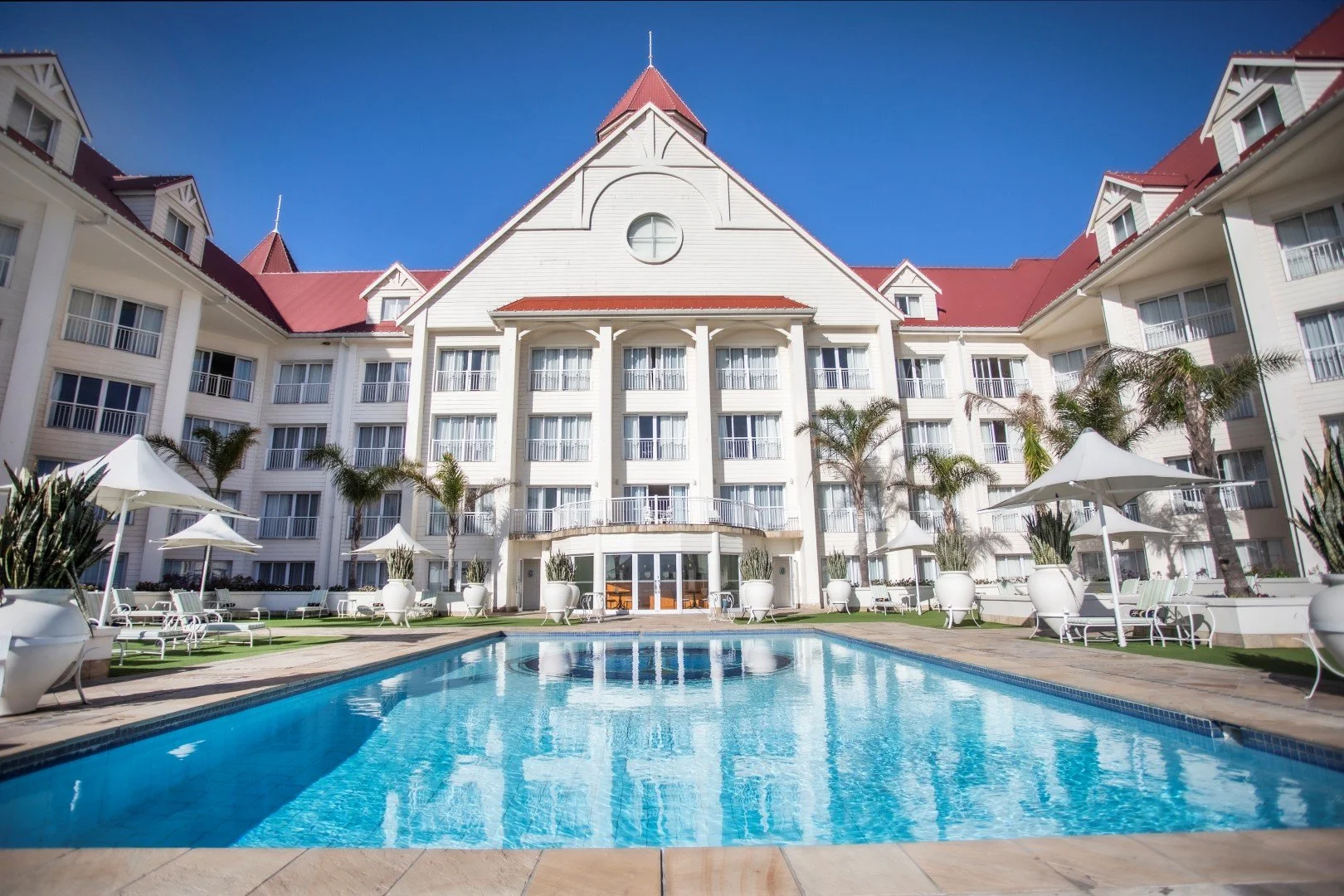 Large white hotel building with a red roof, a central round window, and a swimming pool in front, surrounded by patio furniture, umbrellas, and palm trees, under a clear blue sky.