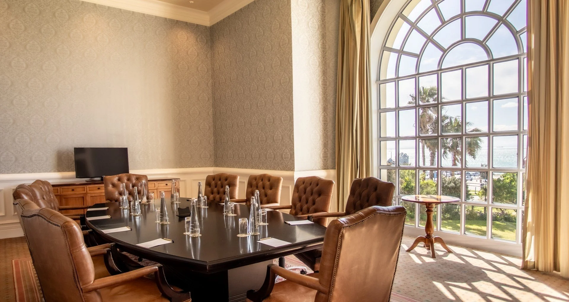 A conference room with a large dark wooden table surrounded by brown leather chairs. The table has water bottles and notepads, with a small TV on a wooden sideboard. A large arched window with beige curtains shows an ocean view with palm trees and a partly cloudy sky.