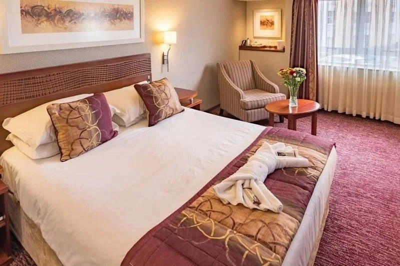 Hotel room with a large bed, a small round table with a vase of flowers, a striped armchair, and a window with curtains.