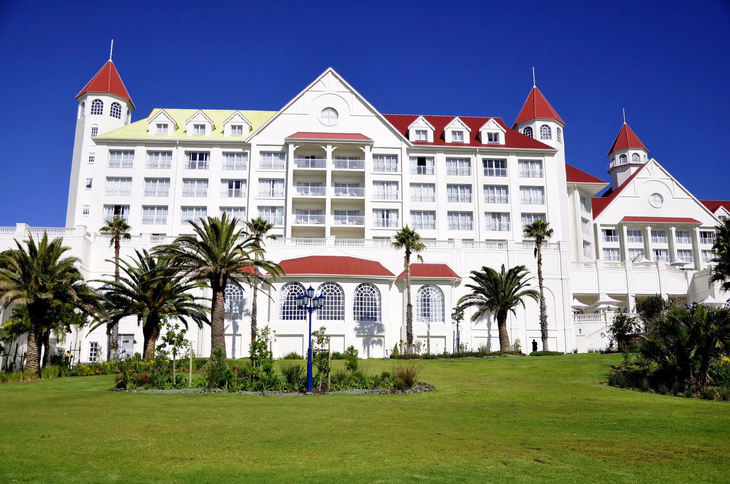 A large white building with red roofs, multiple towers, and balconies. There are palm trees and a grassy area in the foreground, under a clear blue sky.