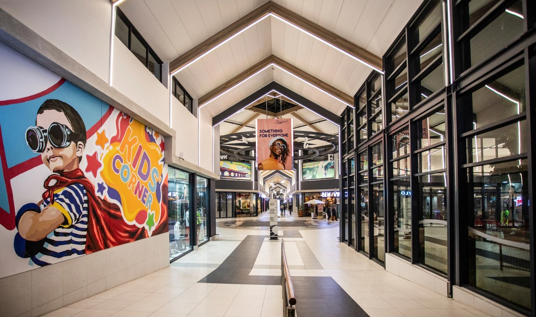 Inside a modern shopping mall with a high, vaulted ceiling and hanging banners. Bright walls feature colorful advertisements, including a large cartoon figure of a child wearing sunglasses and a striped shirt. The corridor is lined with stores, and t