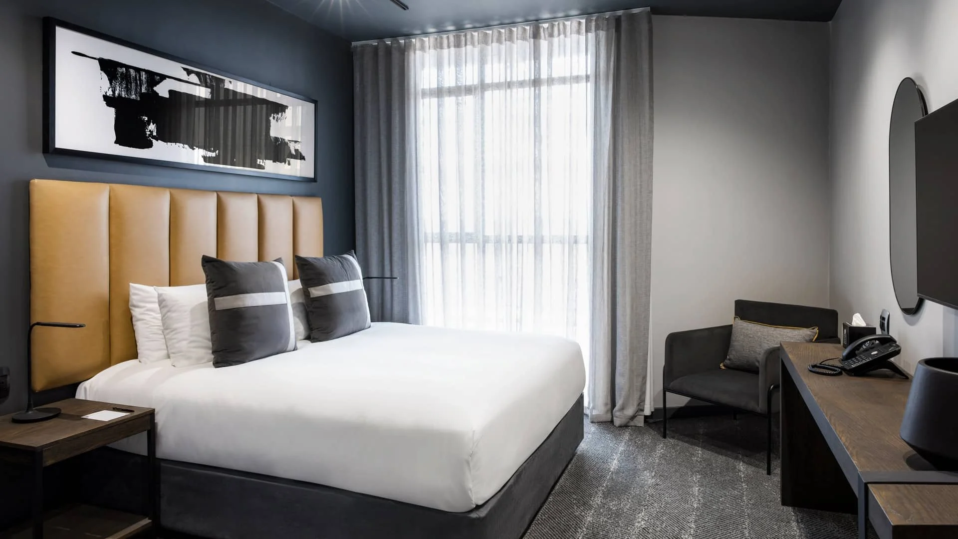 Modern hotel room with a large bed, beige headboard, black and white pillows, nightstand, artwork, window with sheer curtains, small seating area with black chair and cushion, desk with a phone, tissue box, and lamp.