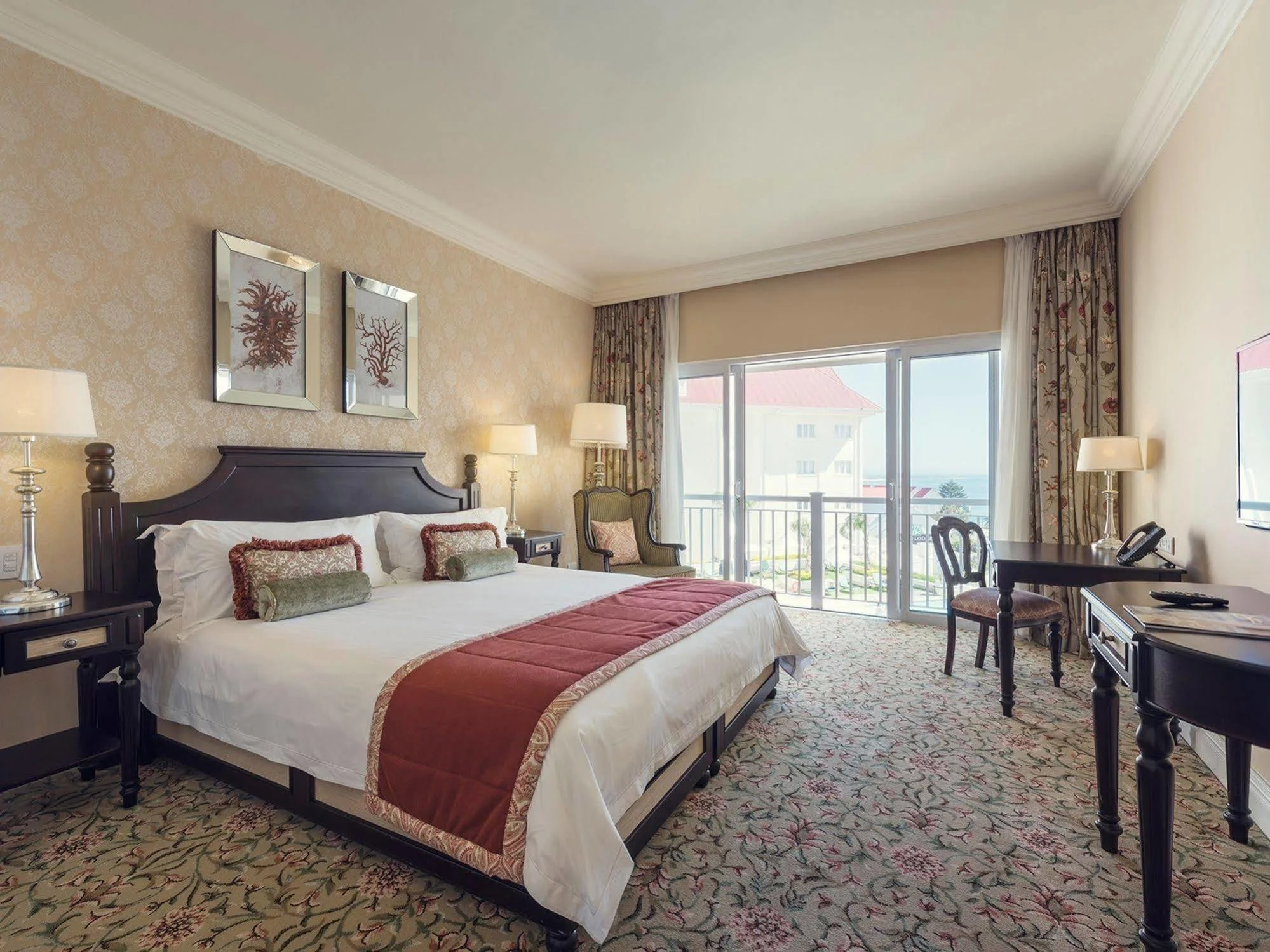 A hotel room with a large bed, two nightstands, table lamps, wall art, a chair, a desk, and a sliding glass door leading to a balcony with a view of neighboring buildings and a distant body of water.