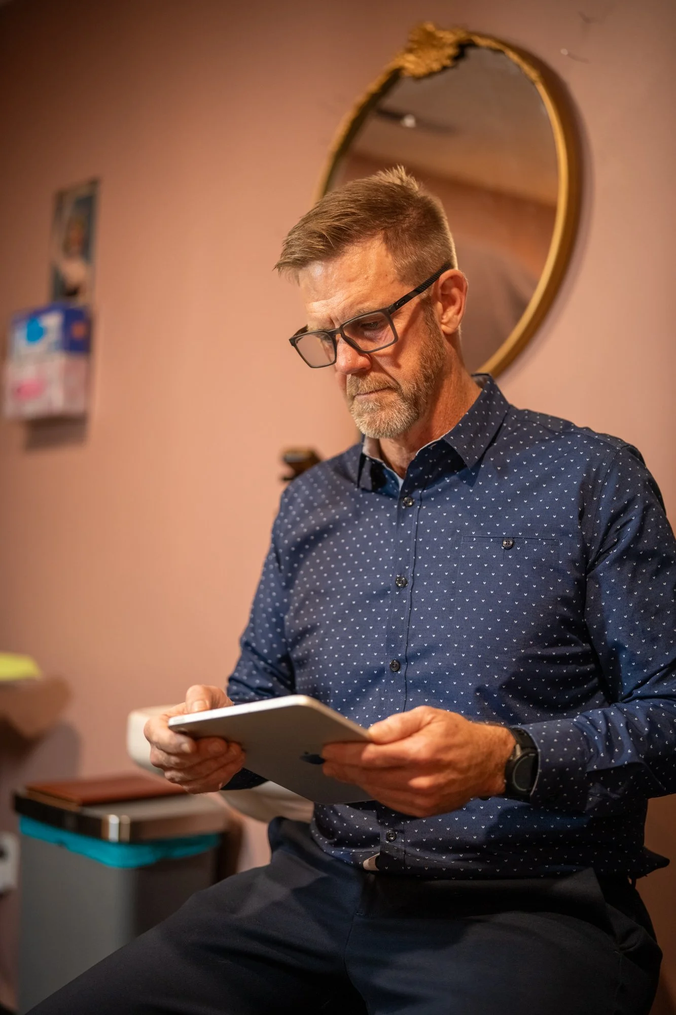 A middle-aged man with glasses and a beard, dressed in a navy blue shirt with white polka dots, is looking at a tablet. He is seated in a room with a pinkish wall and a round mirror behind him.