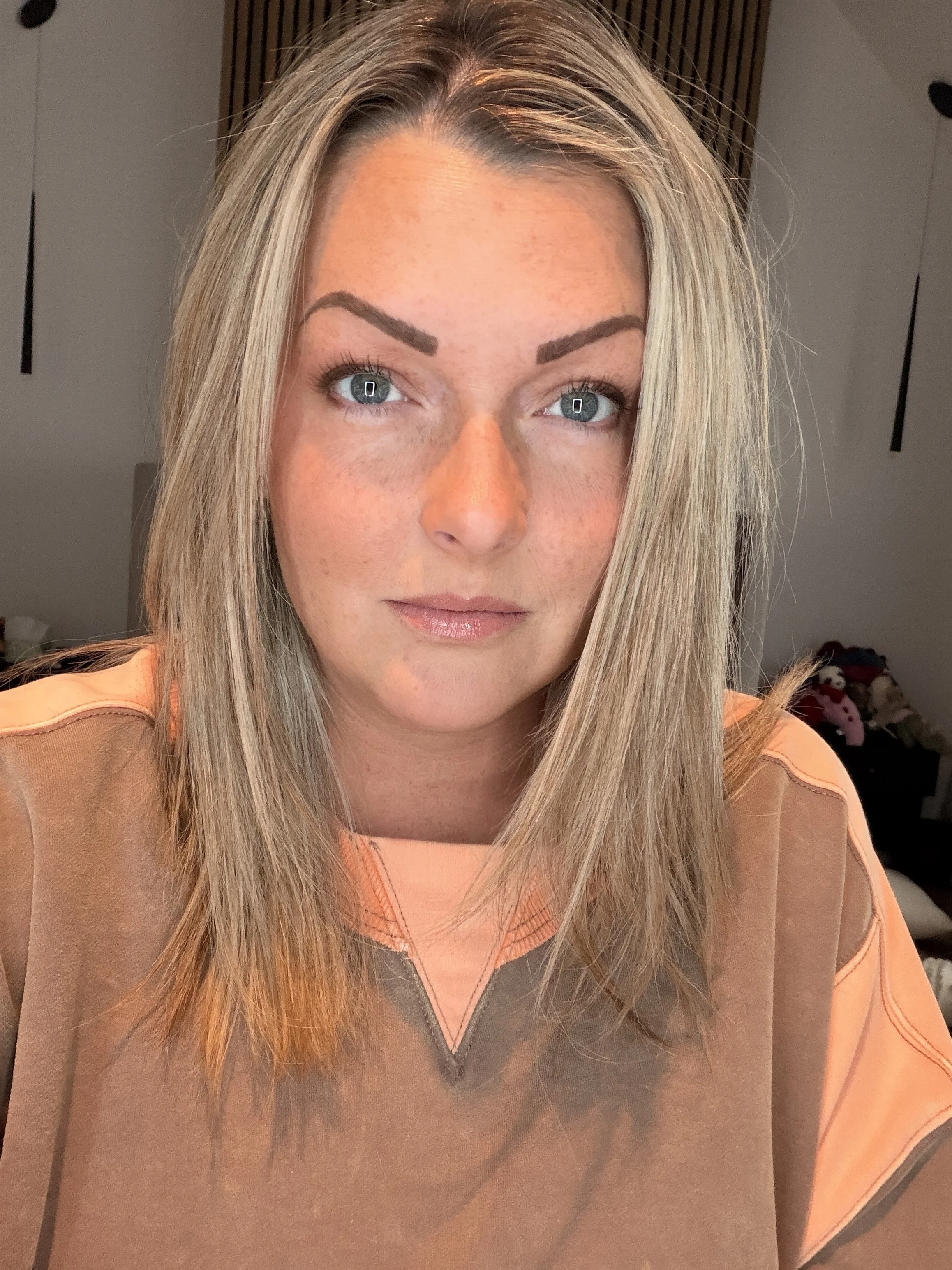 Close-up of a woman with long blonde hair, blue eyes, and makeup, wearing an orange shirt, sitting indoors.