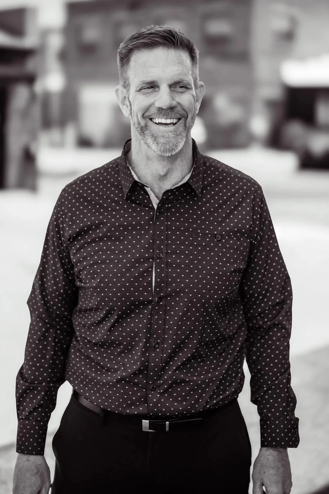 A middle-aged man with a beard and short hair, smiling, wearing a dark shirt with small polka dots, standing outdoors in a blurred background.