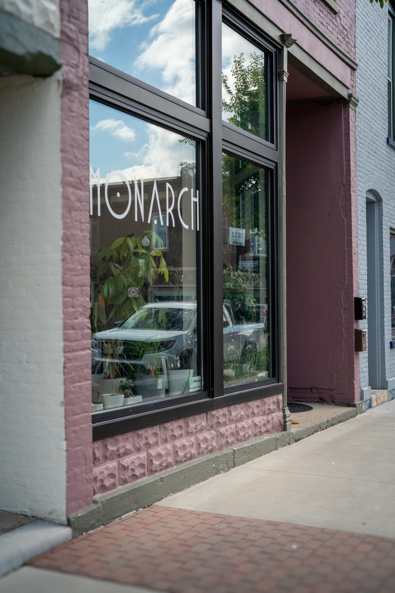 The storefront window of a business with the word 'MOMARCH' displayed on it. The reflection of the cloudy sky and nearby trees can be seen in the glass. Inside, there are potted plants visible through the window.