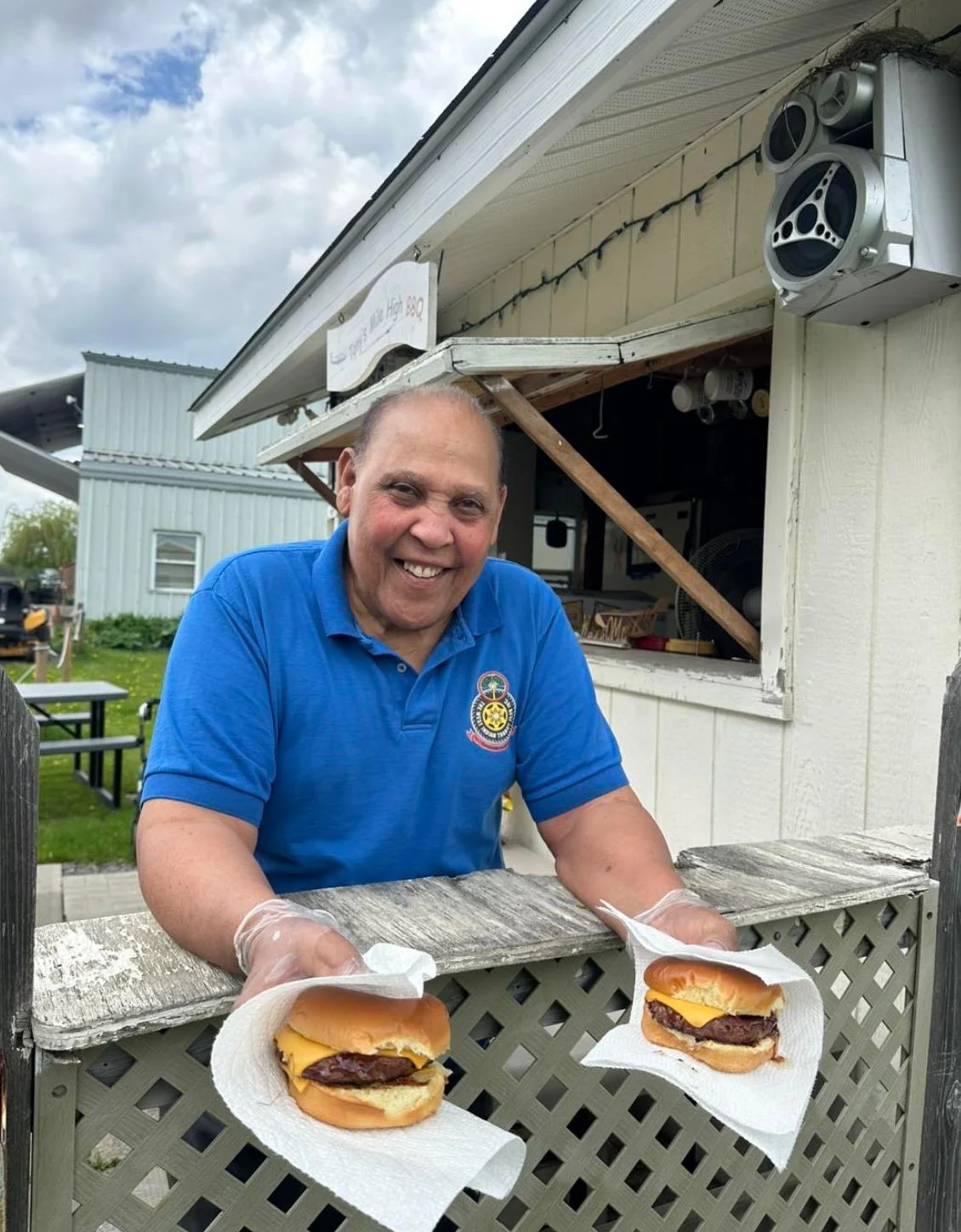 A smiling man wearing a blue polo shirt and gloves stands behind a lattice fence, holding two cheeseburgers wrapped in paper towels. There is a small white building or food stand behind him with an open window and a speaker mounted on the corner.