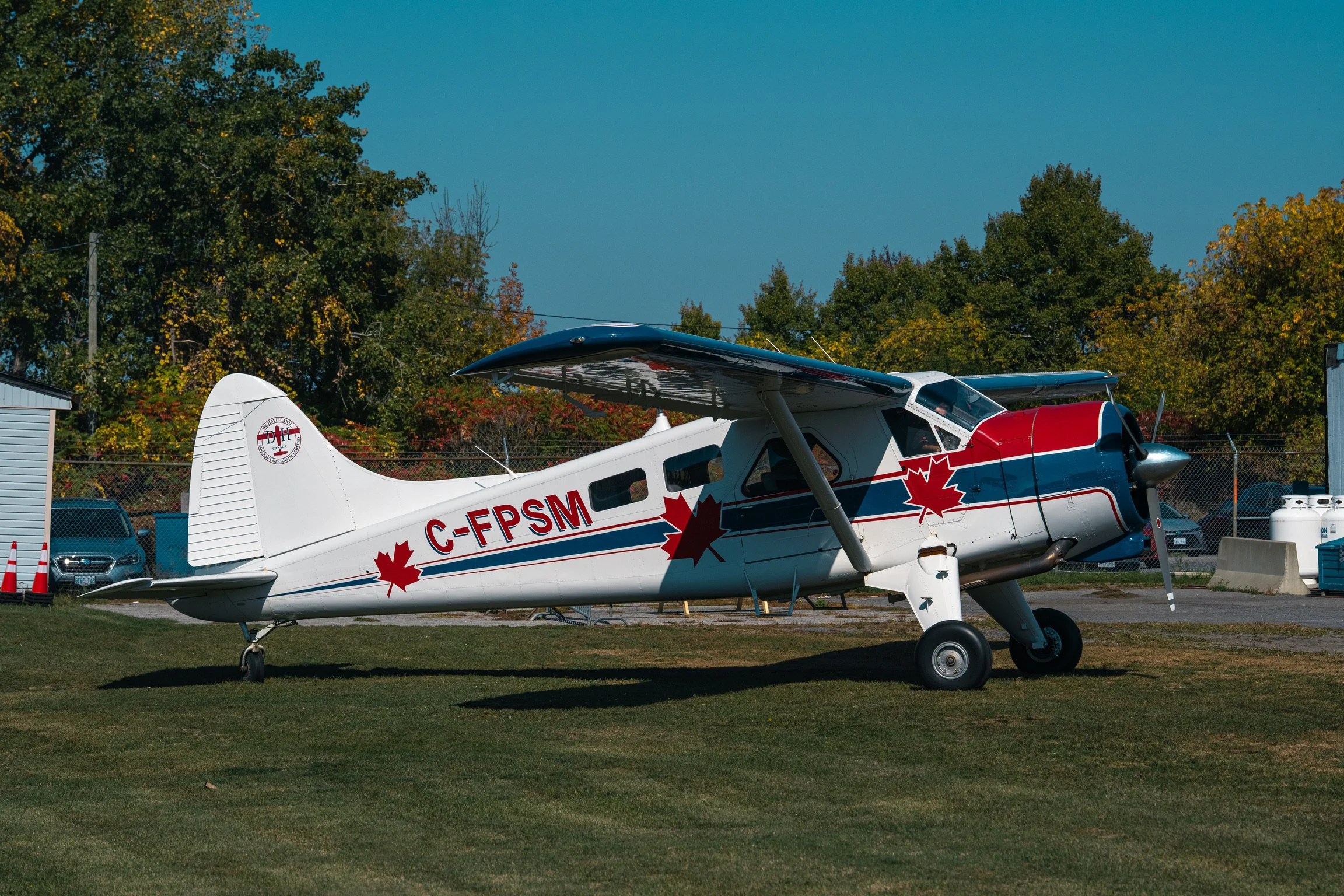 Small vintage propeller airplane with a white body and red, blue, and gray accents, parked on a grassy area near trees and vehicles under a clear blue sky.