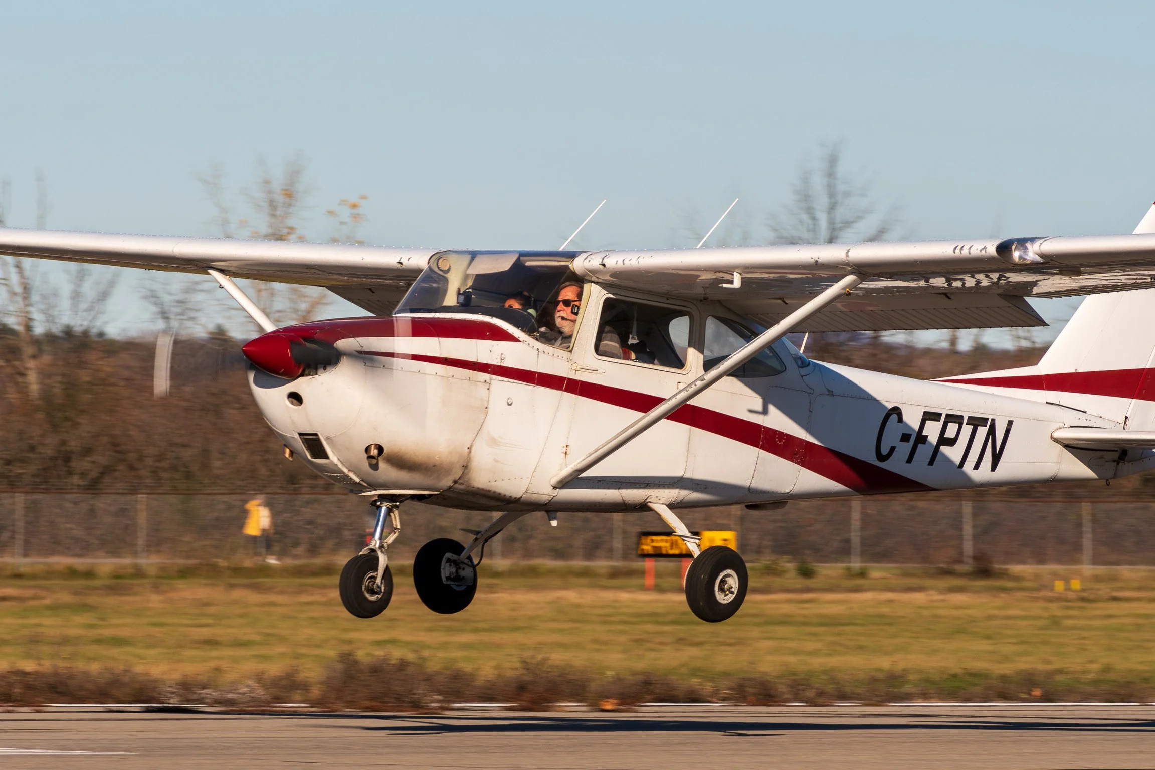 A small white airplane with red stripes is taking off from a runway, with the pilot visible through the cockpit window, and the sky is clear with some distant trees.