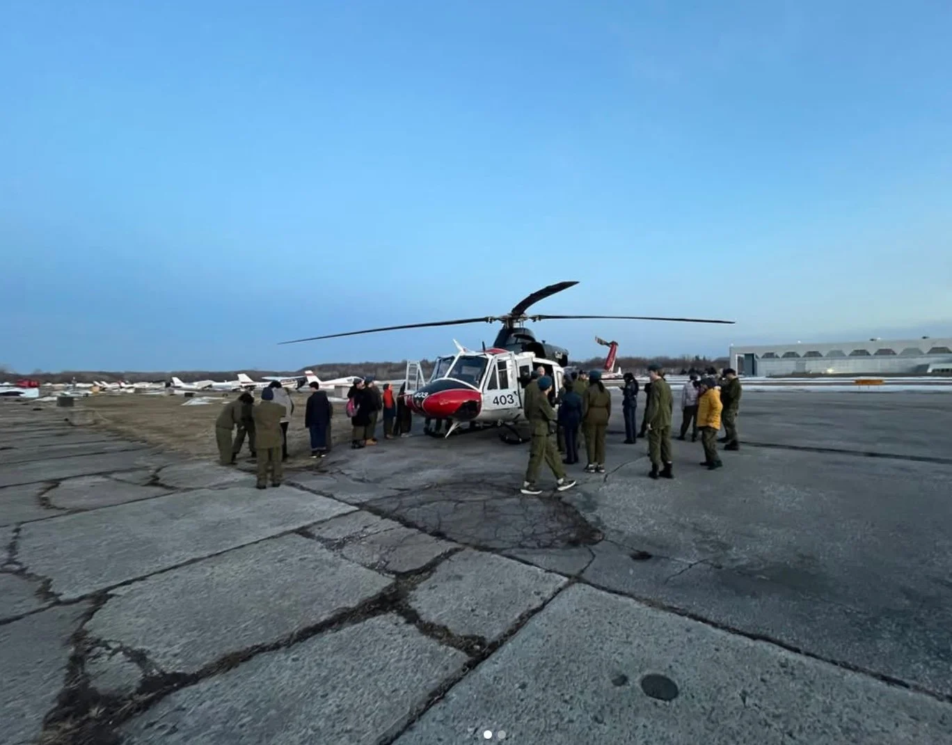 Group of people, including military personnel, gathered around a helicopter on a cracked tarmac at an airport, with several small aircraft and airport facilities in the background under a clear blue sky.