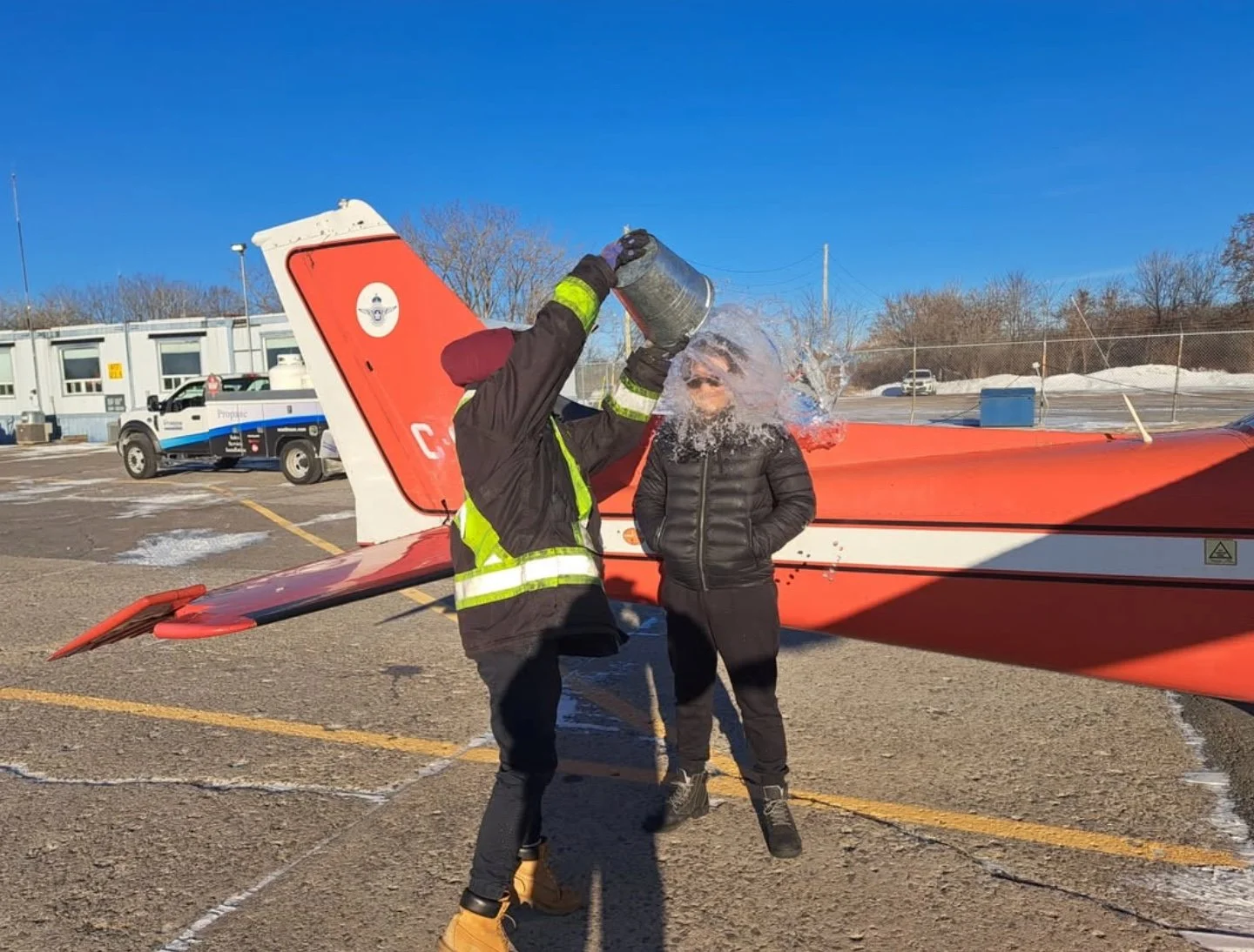 Firefighter pouring water over a woman’s head in a parking lot with a small red airplane behind them and clear blue sky.