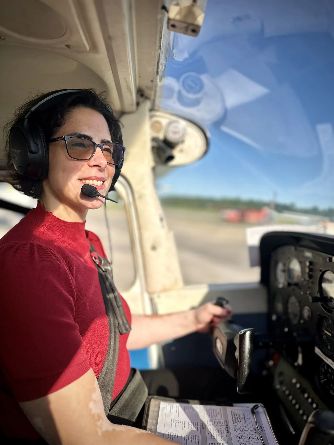 A woman with short dark hair, wearing glasses and a red shirt, sitting in the cockpit of a small airplane, wearing a headset and smiling.