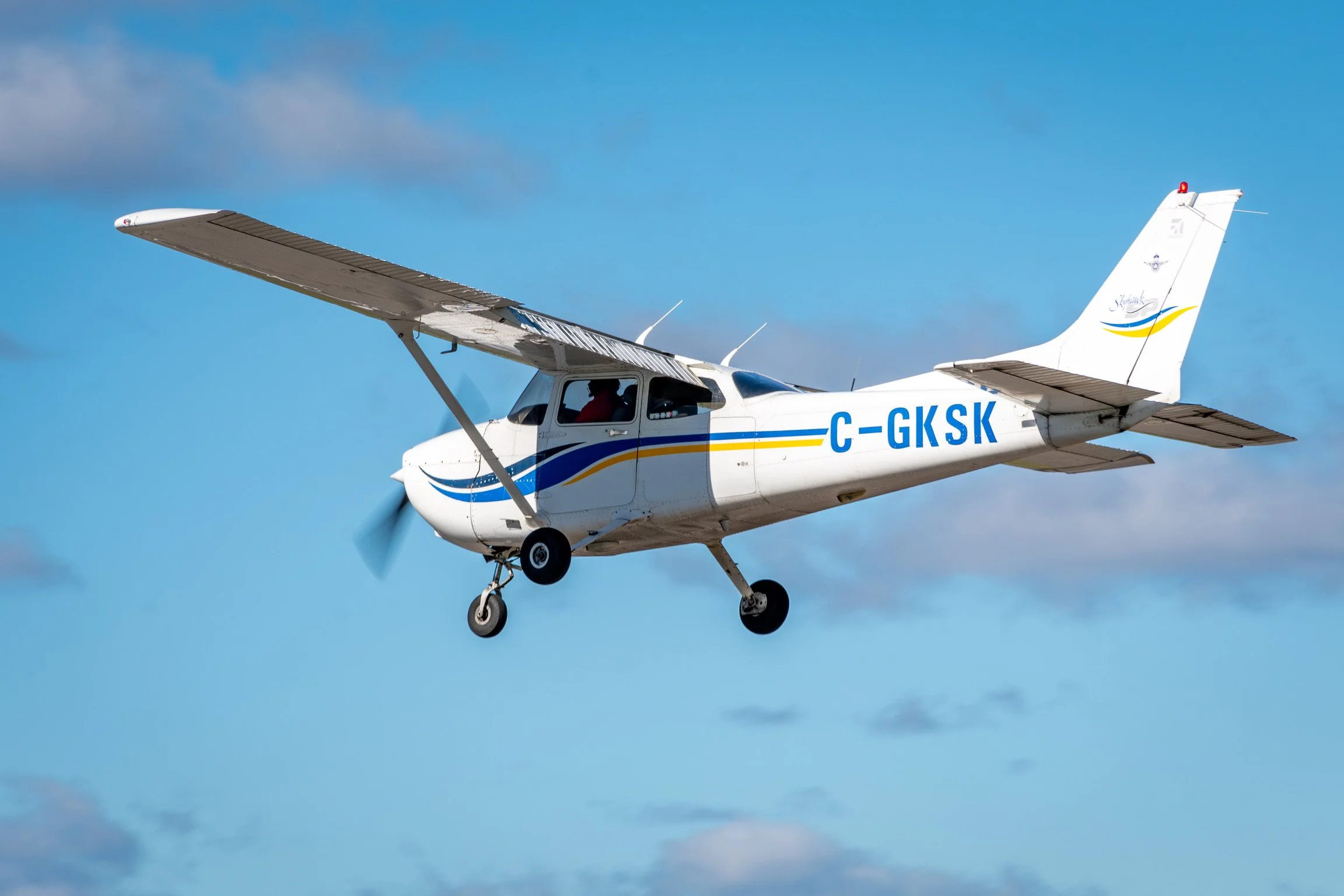 Small white airplane with blue and yellow stripe flying in blue sky with clouds.