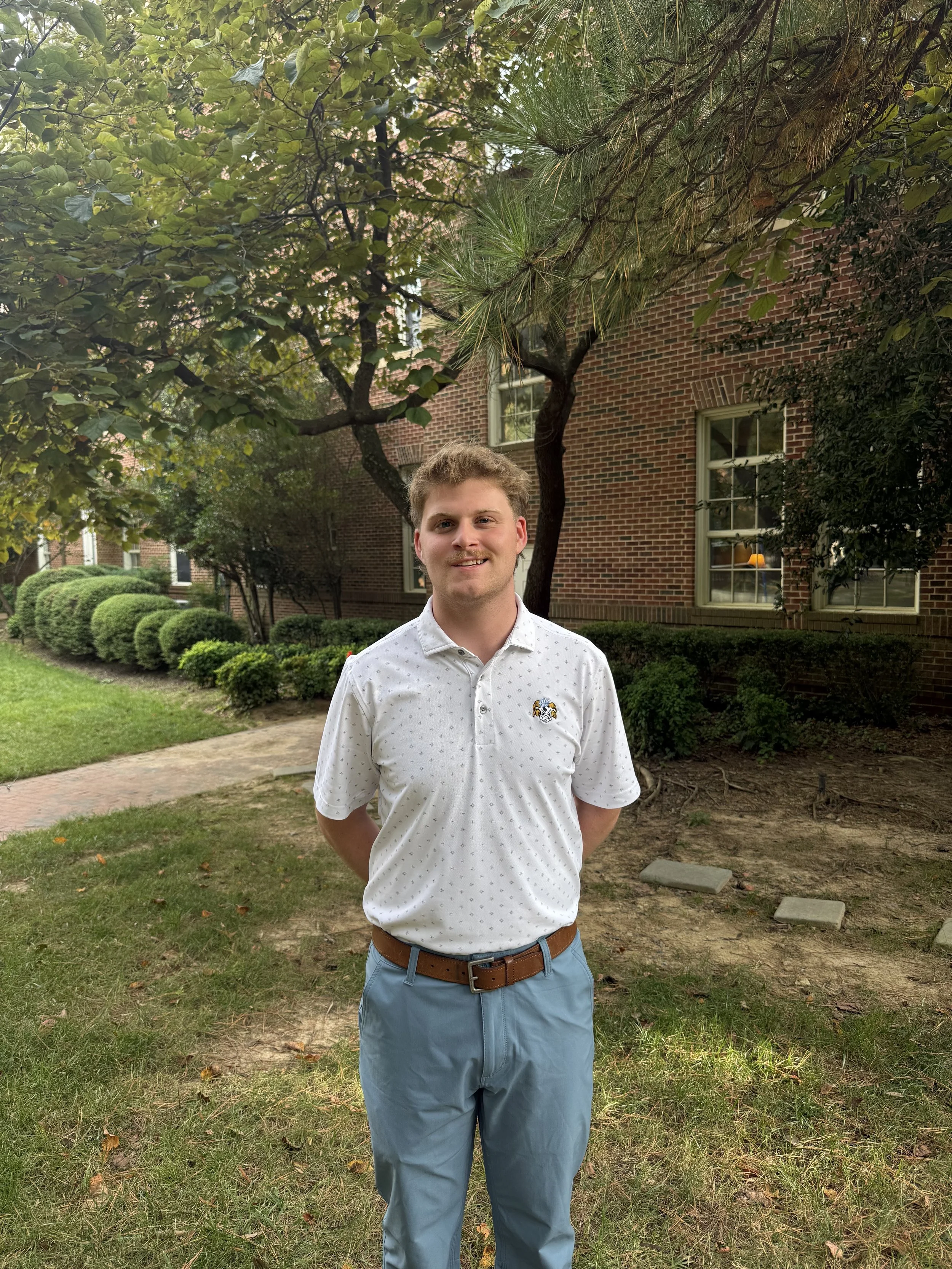 A young man with light brown hair, wearing a white shirt with a UNC logo on it and light blue pants, standing outdoors on a grassy area with trees and a brick building in the background.
