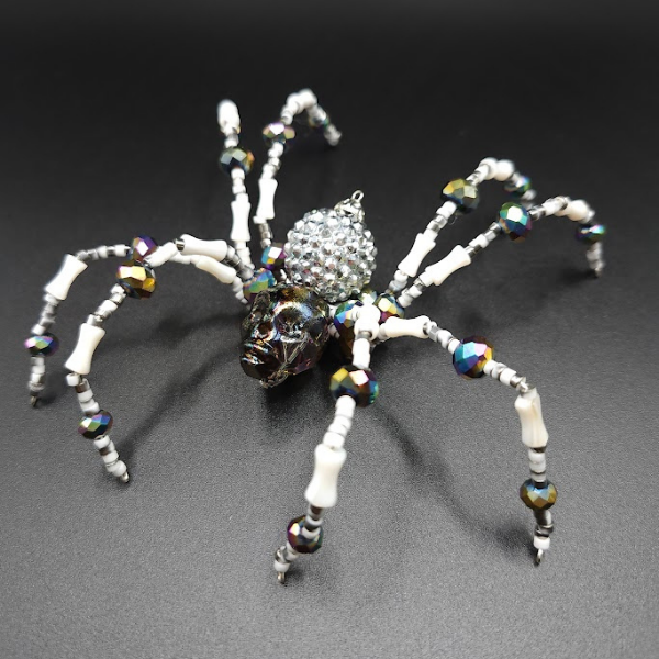 Beaded spider ornament with a skull-shaped body and a rhinestone-covered abdomen on a black background.