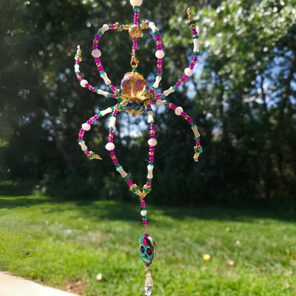 Colorful beaded spider ornament with a skull charm hanging from its abdomen, suspended outdoors with trees and grass in the background.