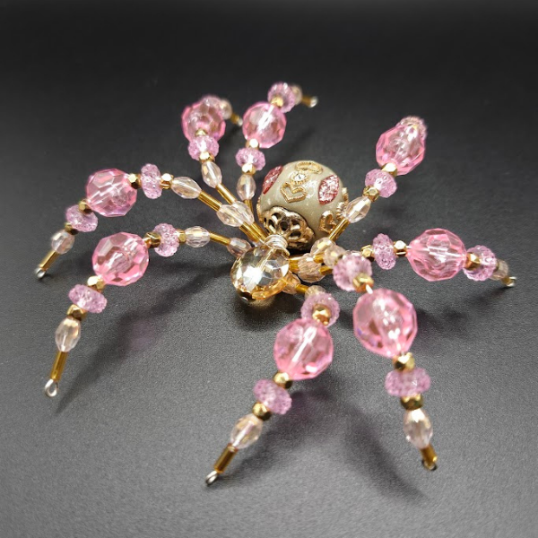 Pink and clear beaded spider-shaped jewelry with a decorative skull at its center, lying on a black surface.