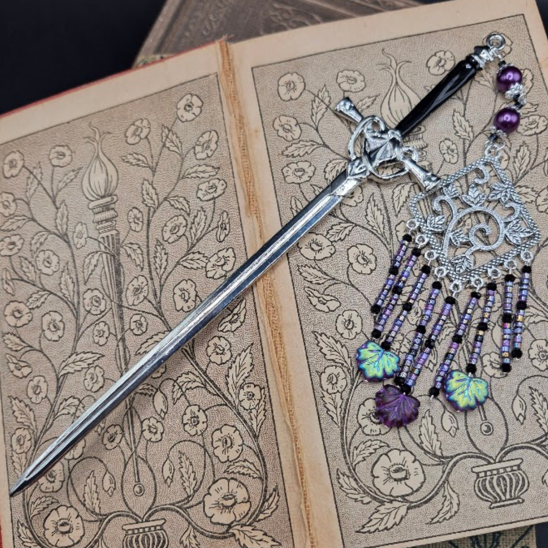 An open vintage book with floral illustrations, a decorative dagger, and a silver and purple beaded charm with purple leaf charms placed on it.