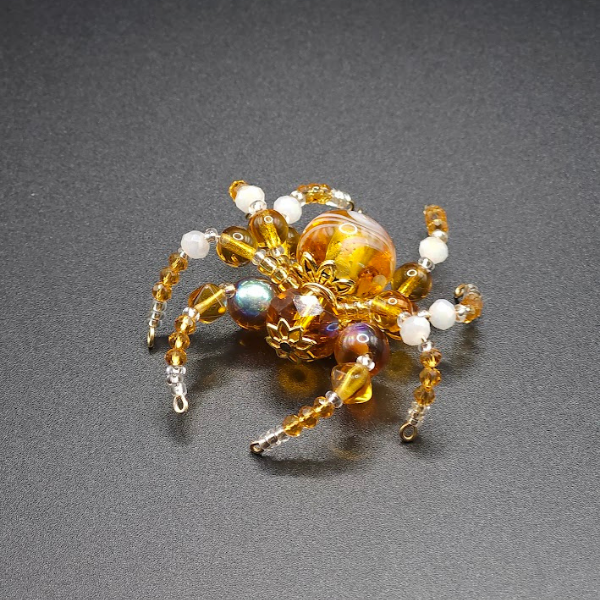 A decorative spider made of various beads, including gold, white, pearl, and amber-colored beads, arranged to resemble a spider with beads as its body and legs, on a black surface.