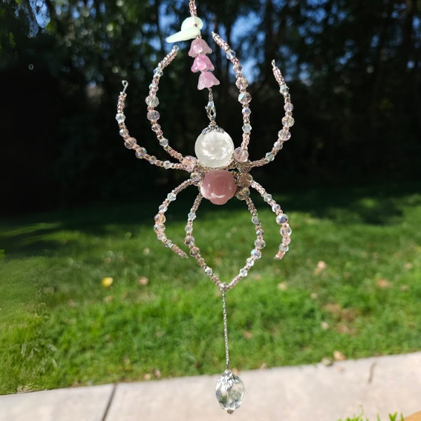 Beaded ornament shaped like a spider with pink, white, and clear beads, hanging outdoors with grass and trees in the background.