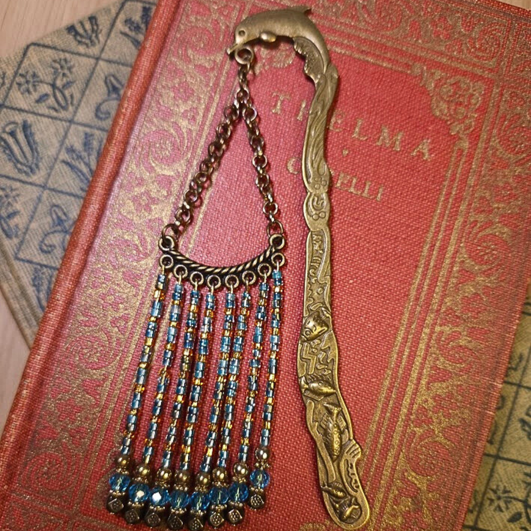 A vintage gold-tone spade-shaped bookmark with an attached blue and gold beaded chain on a red textured book with gold accents.