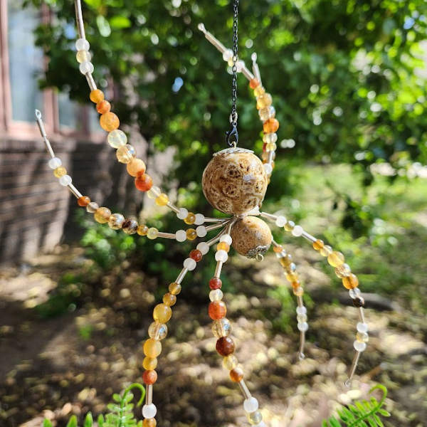 A spider made of beads and stones, with a natural stone at the center, hanging outdoors with greenery in the background.