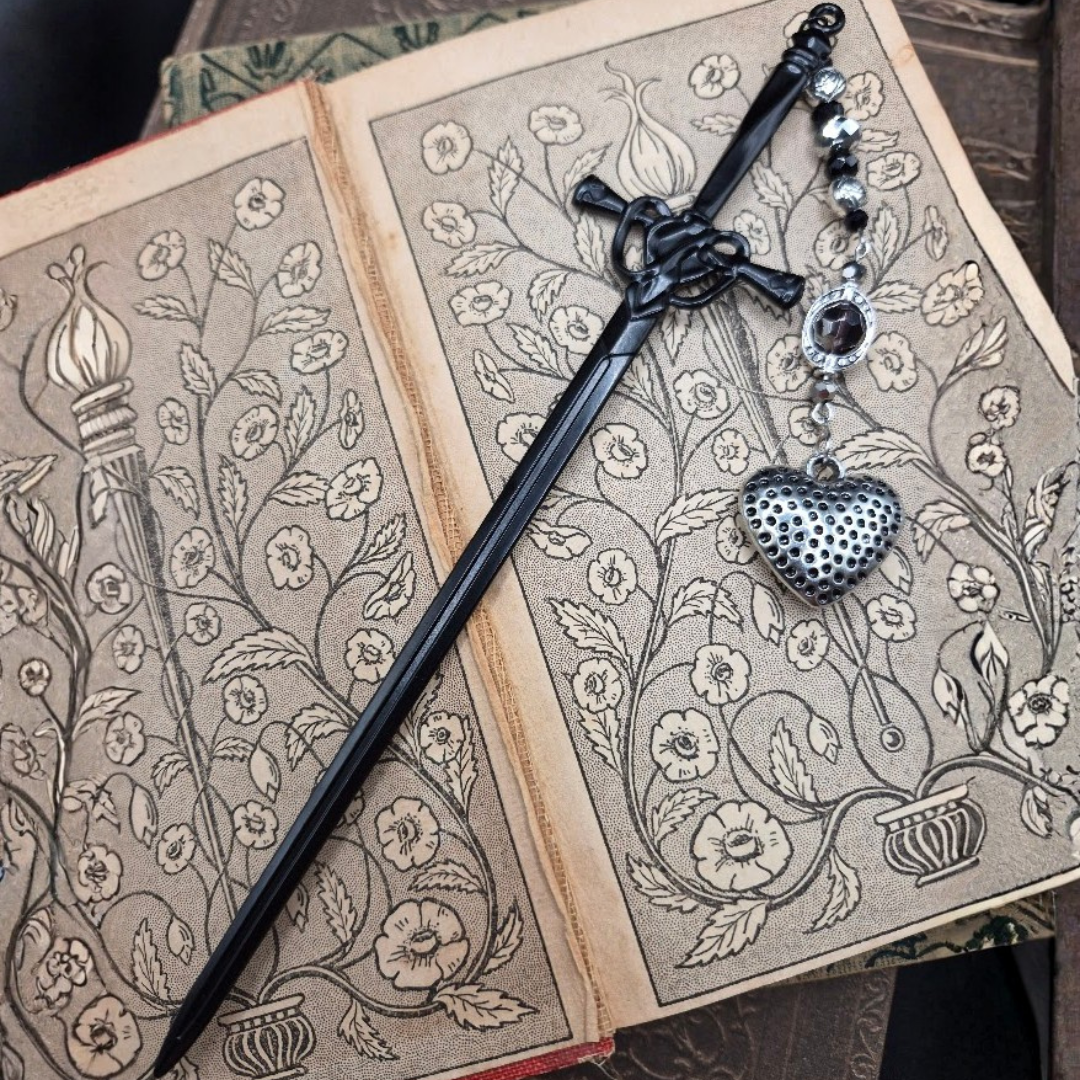 An open book with intricate floral illustrations, a black sword bookmark with beads and a black heart-shaped pendant on top.