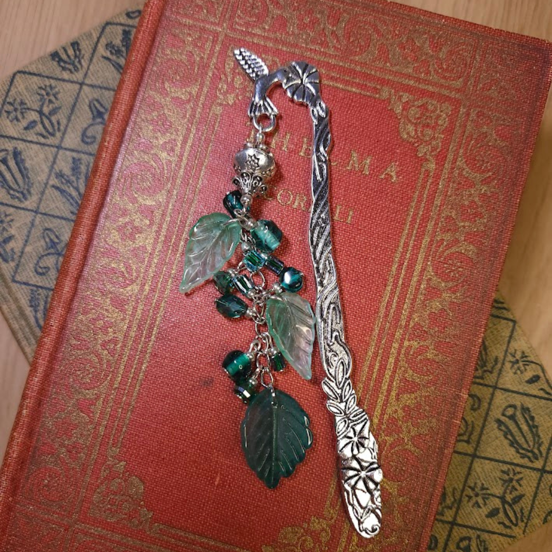 A vintage silver bookmark with intricate floral engravings and a charm featuring green glass leaves and beads, resting on a red textured book cover.