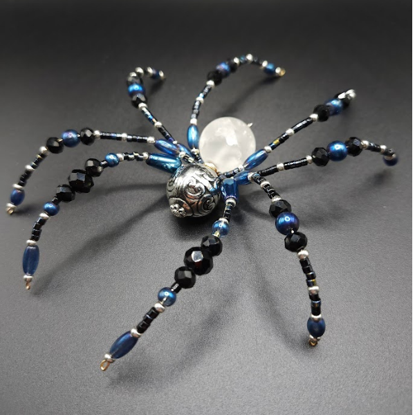 A beaded spider-shaped ornament with black and blue beads, featuring a large silver bead and faceted black beads, on a black background.