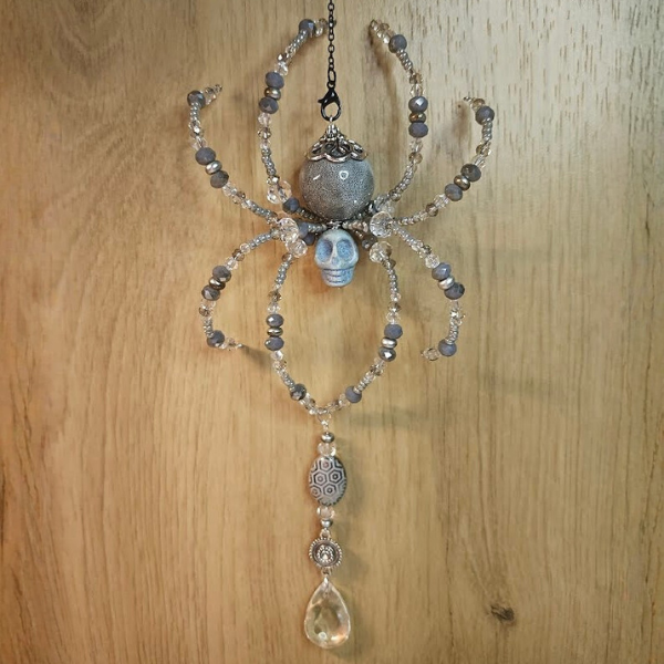 Decorative spider-shaped ornament with beaded legs, a skull center, and a hanging teardrop-shaped gemstone, hanging on a wooden surface.