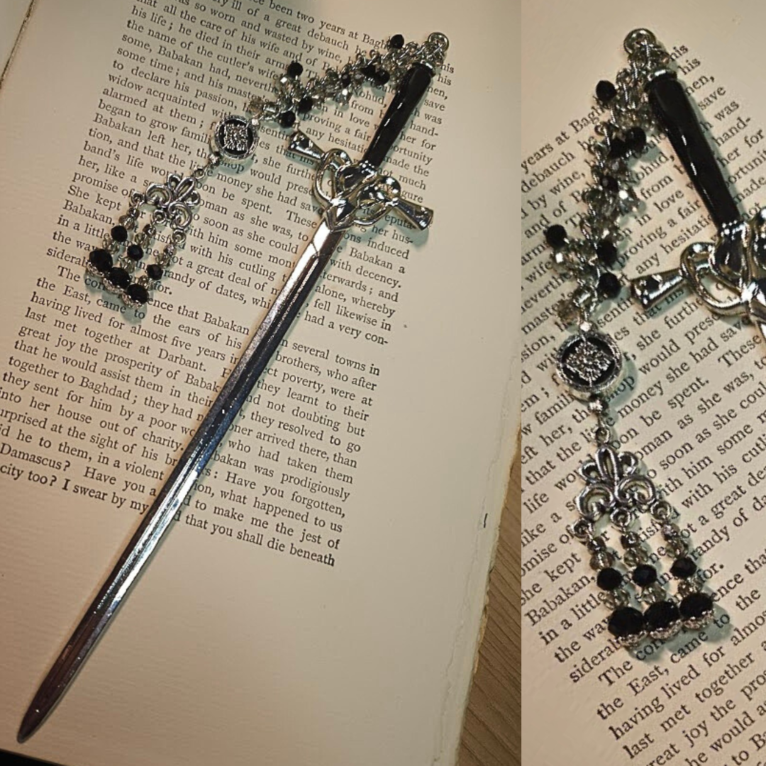 Silver ornate book mark with black and clear gemstones and a cross charm, resting on an open book with printed text.
