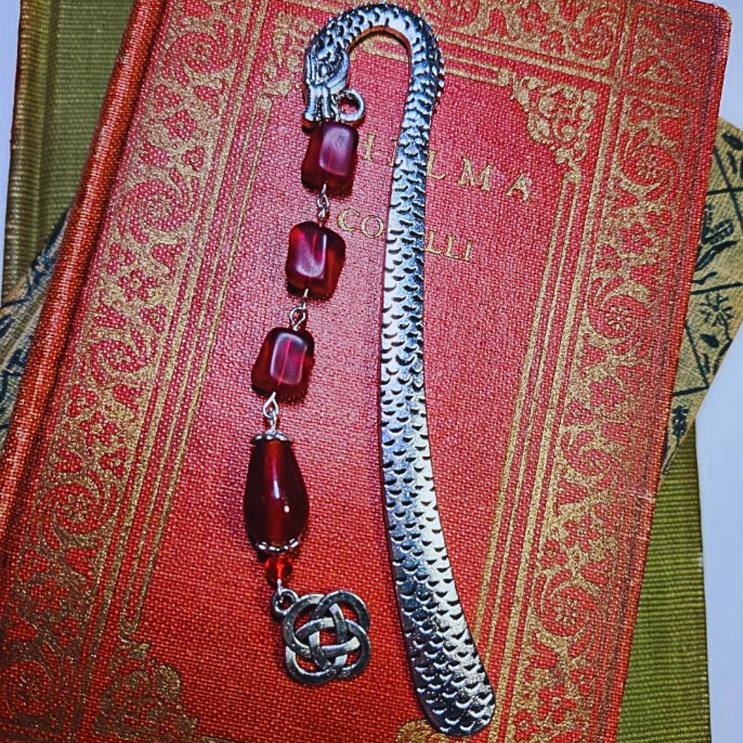 An ornate book with gold and red cover next to a silver bookmark with red beads and a Celtic knot charm.