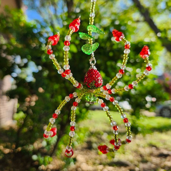 Beaded spider ornament with red, green, and clear beads, hanging outdoors with trees and a sunny sky in the background.