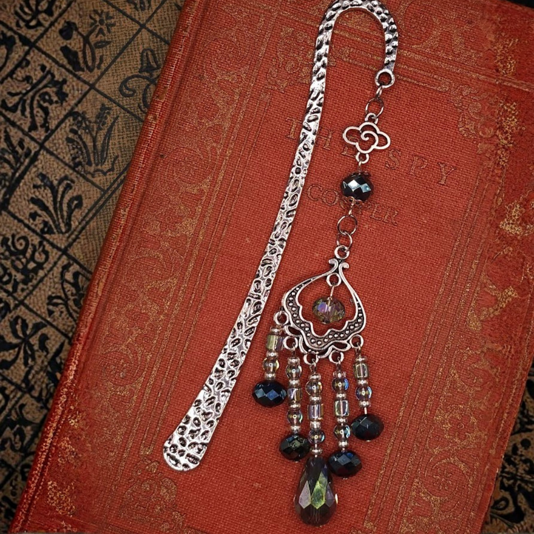 A vintage silver bookmark with black beads and a decorative pendant, displayed on a red textured cloth, with an ornate black and gold patterned book cover partially visible.