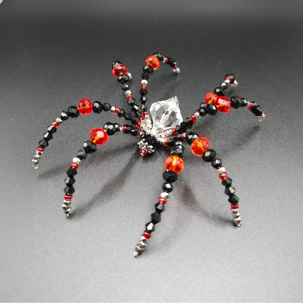 A spider-shaped ornament made of black, red, and silver beads with a large clear bead as its body, set against a black background.