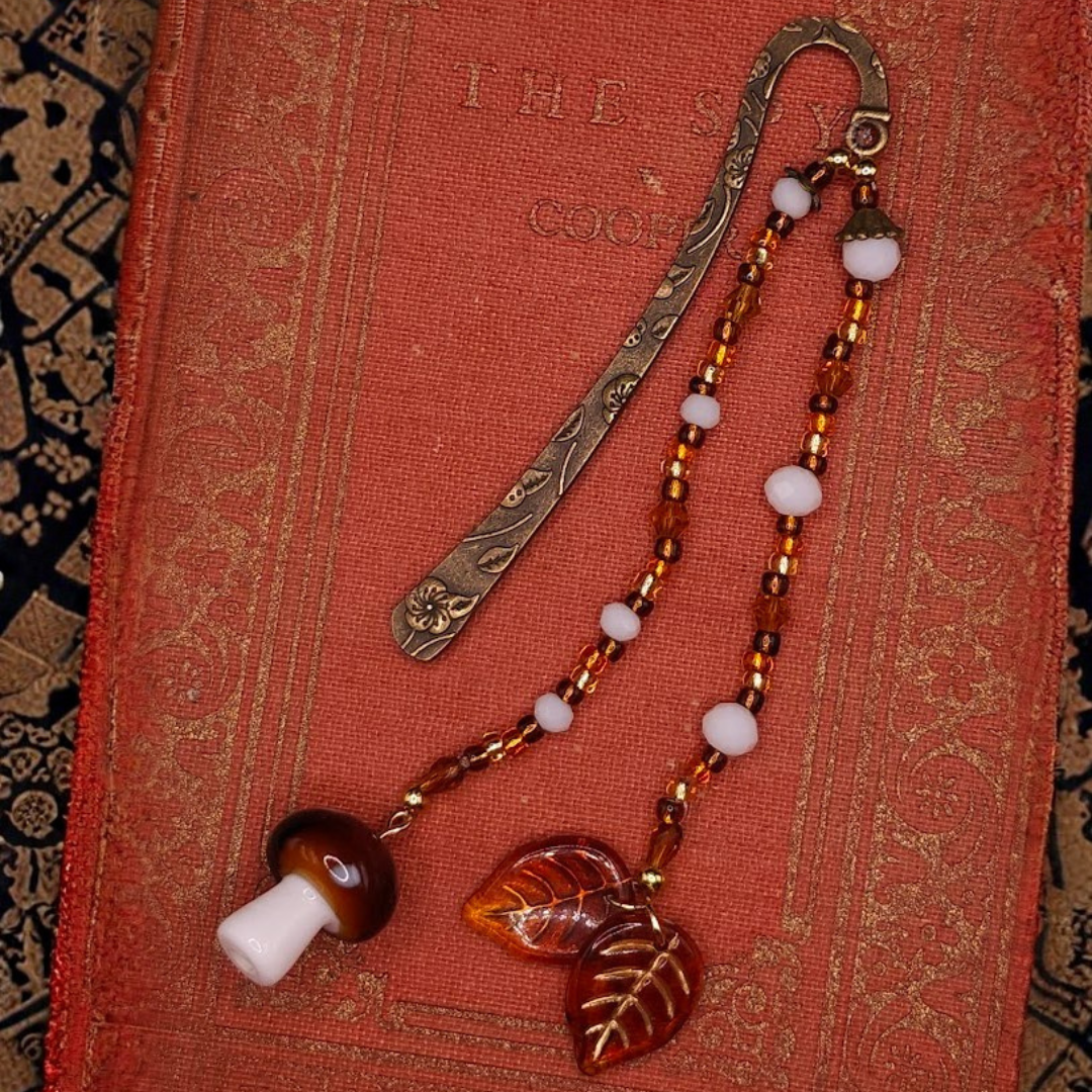 An ornate, vintage-style book with a decorative metal bookmark and a beaded necklace with white, amber, and brown beads, featuring a mushroom and leaf-shaped pendant, laid on top.