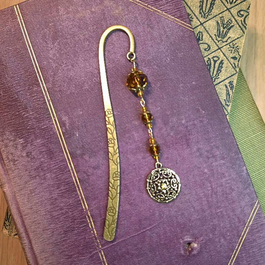 A decorative bookmark with a metal charm and amber-colored beads resting on a purple book.