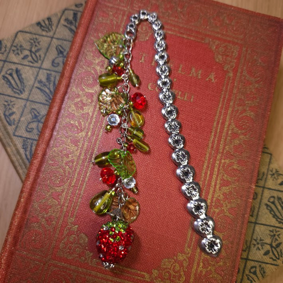 A colorful charm bookmark with red, green, and strawberry beads, placed on a red ornate book with gold lettering, next to another book with a decorative blue and beige cover.