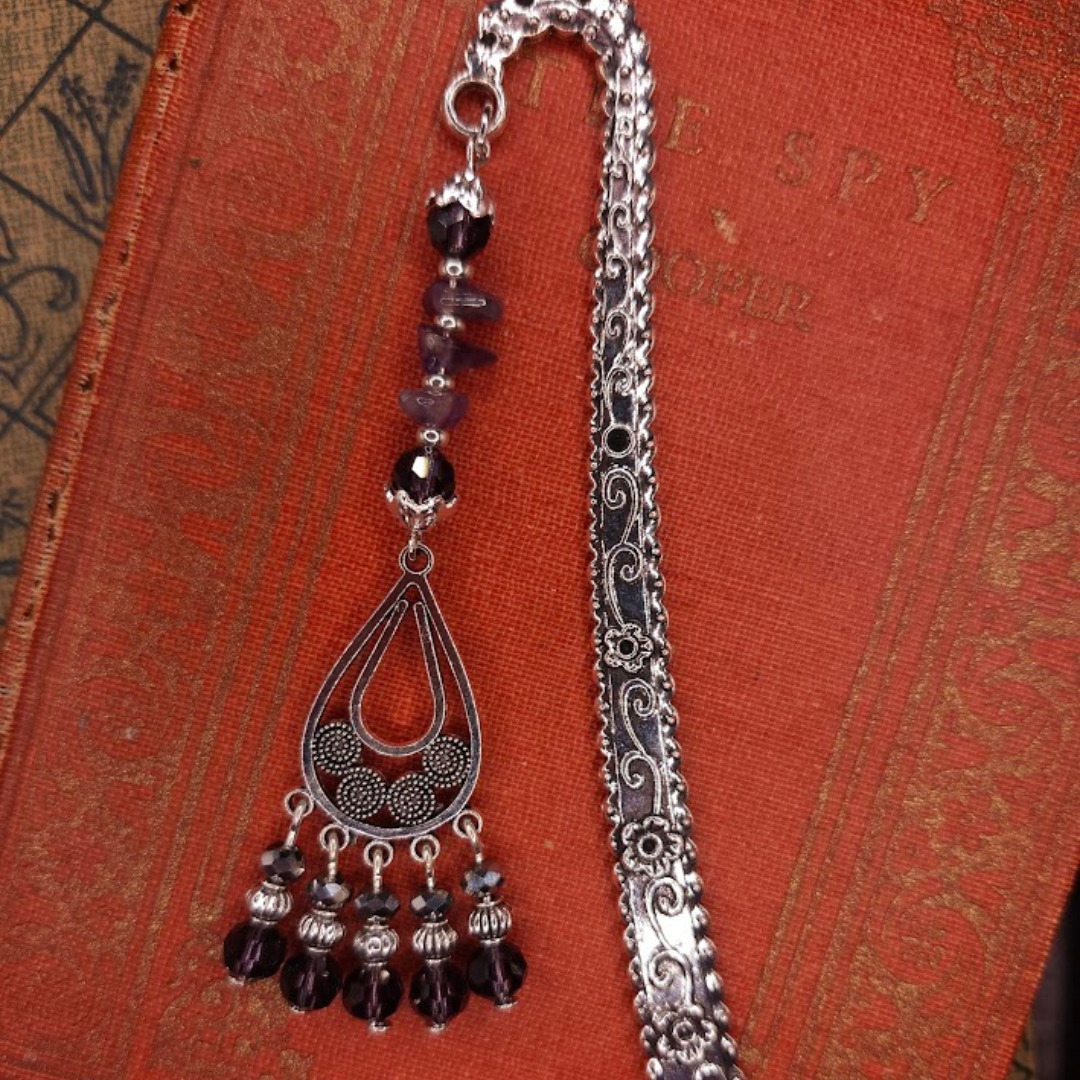 A silver bookmark with intricate designs and dangling beads resting on an orange textured surface with gold patterns.