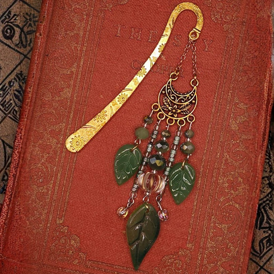 Decorative bookmark with green leaves, beads, and a large leaf pendant, placed on a red textured surface.