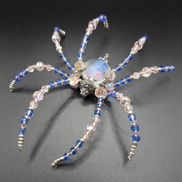 A jewelry piece resembling a spider, made with blue, pink, and clear beads, with a central blue bead and decorative silver elements, placed on a dark background.