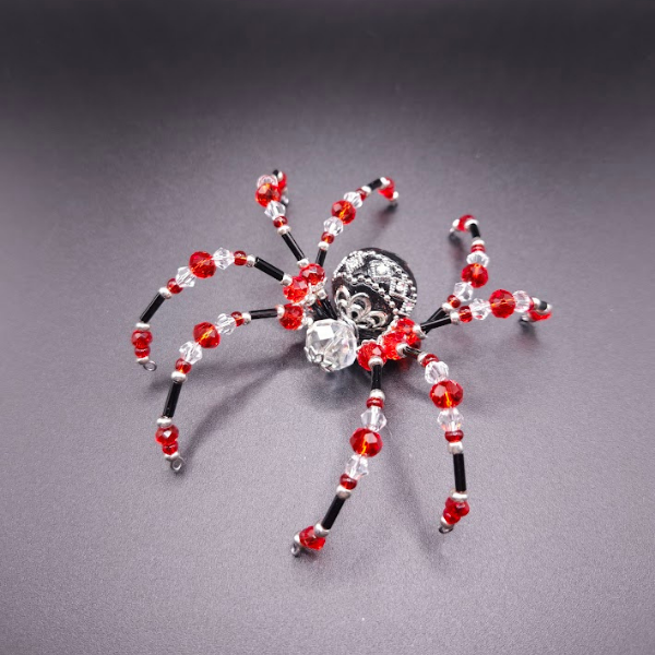 A decorative spider made of red, black, and clear beads with a silver skull-shaped bead as the body, placed on a gray surface.