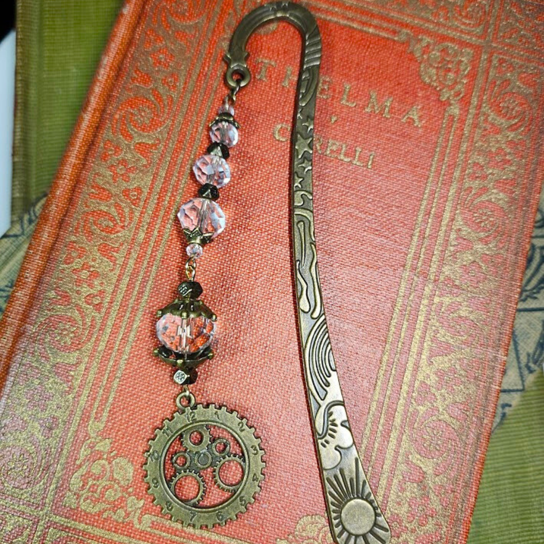A vintage bookmark with a steampunk gear pendant and crystal beads, resting on a red textured book cover.